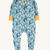 The Frugi Lovely babygrow in a blue penguin waddle design. A hand illustrated style, all over penguin print on a blue background with yellow gold coloured fold-able cuffs on the arms and yellow gold coloured binding on the neck. 