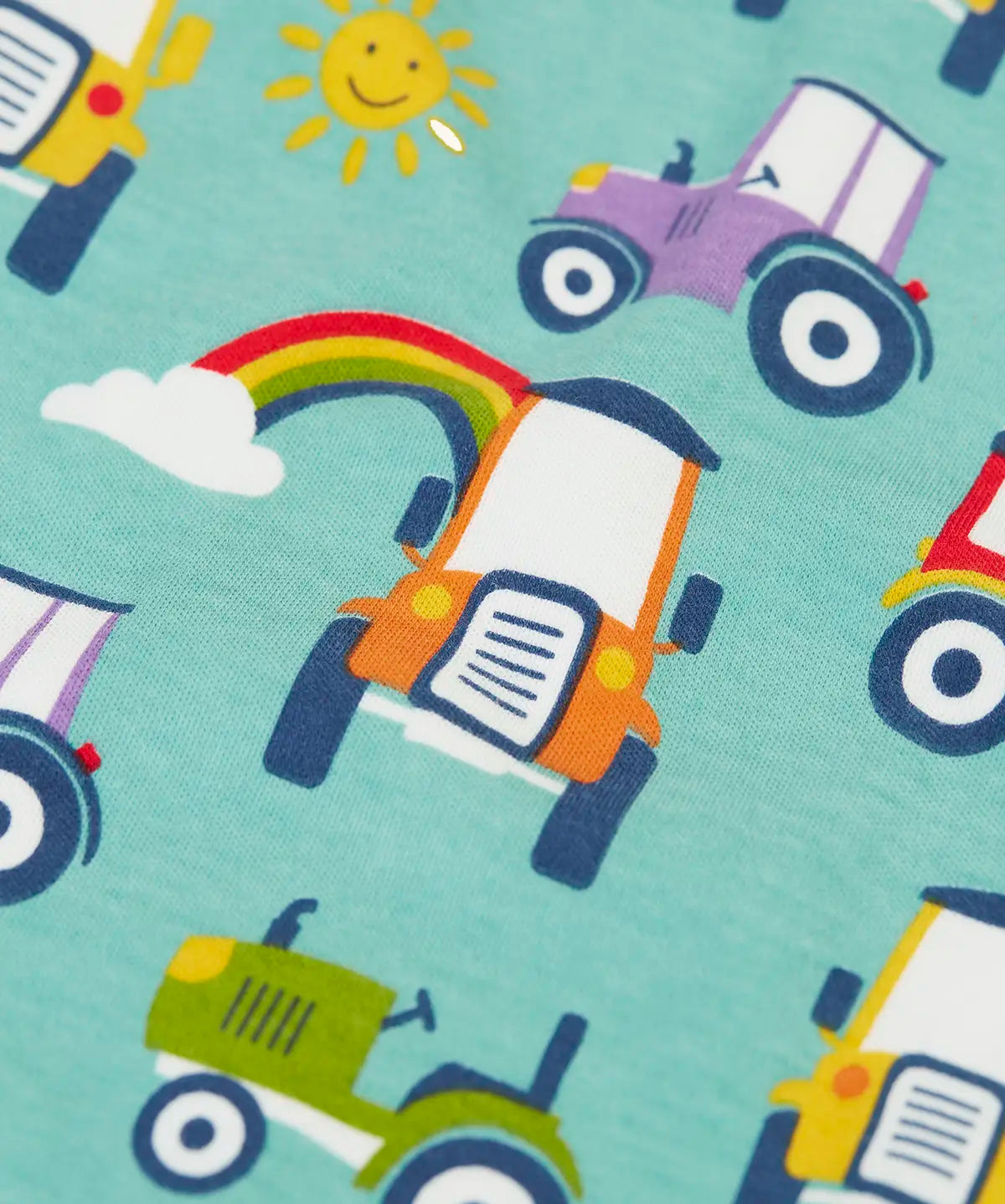 Close up of the colourful tractors on the Frugi organic cotton lovely babygrow sleepsuit with an all over terrific tractors print on a blue background. 