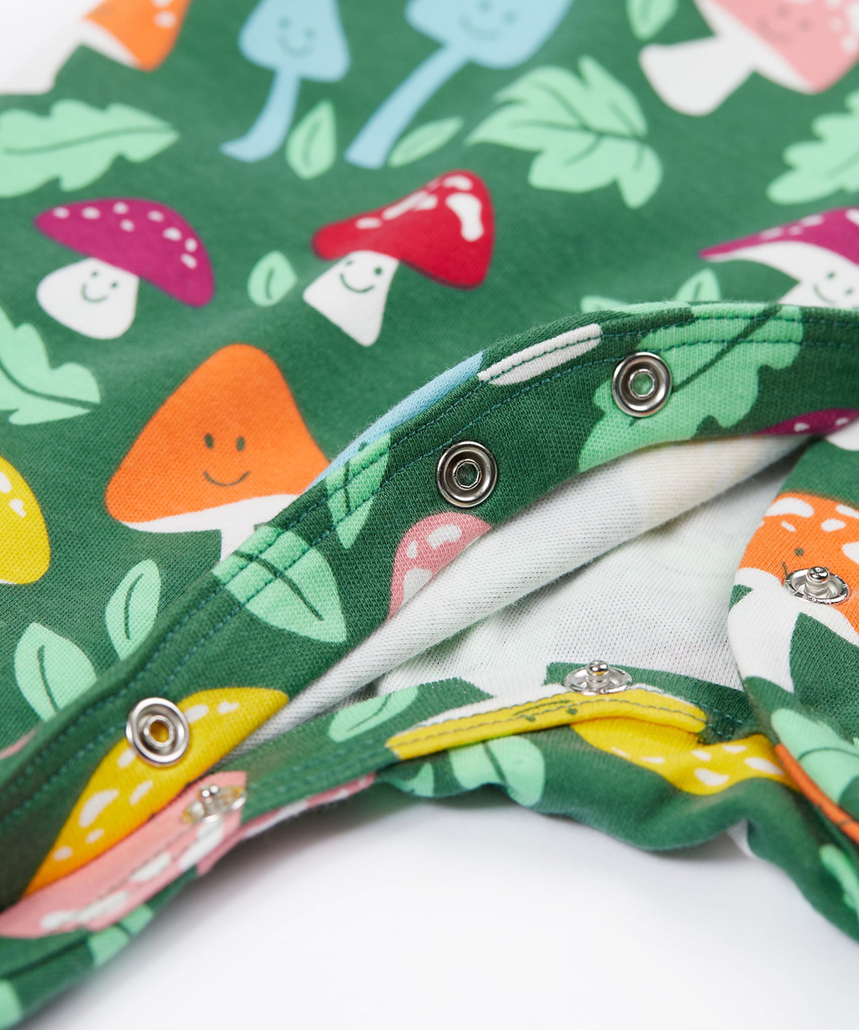 A close up of the popper closure on the Frugi Lovely Babygrow with a fun guys design. The green coloured organic cotton babygrow has multi coloured mushrooms with happy faces.