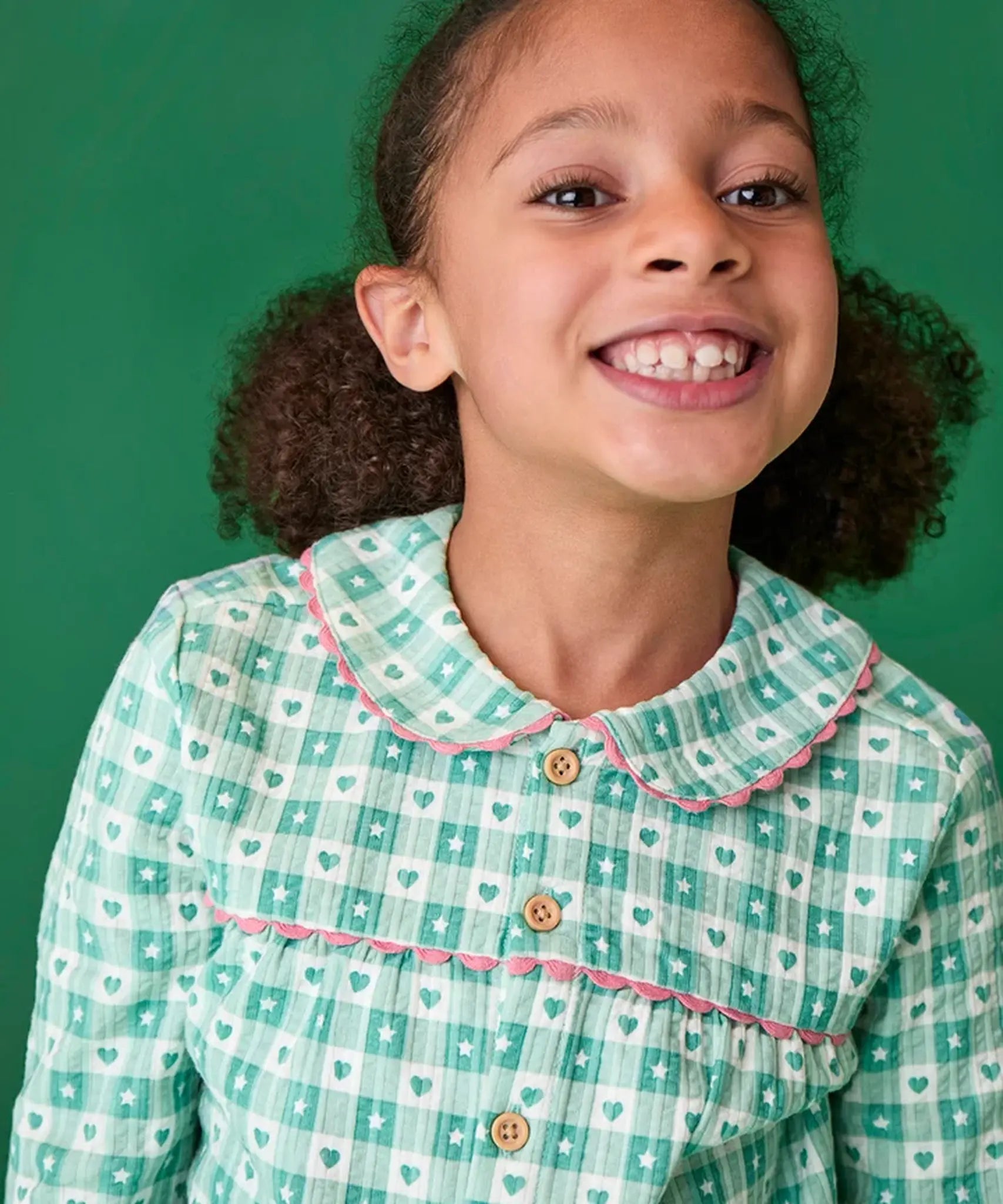 Close up of hild wearing the Frugi magic nights organic cotton hayley traditional pyjamas. Light green and white check style with hearts and stars, pink frill around the collar and across the chest.