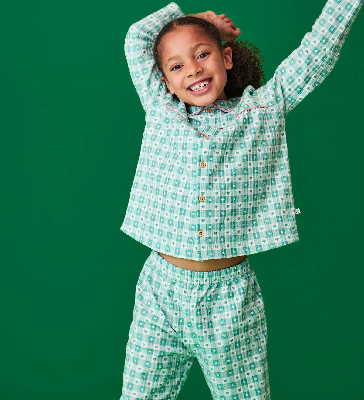 Child wearing the Frugi magic nights organic cotton hayley traditional pyjamas. Light green and white check style with hearts and stars. Pink frill around the collar and across the chest.