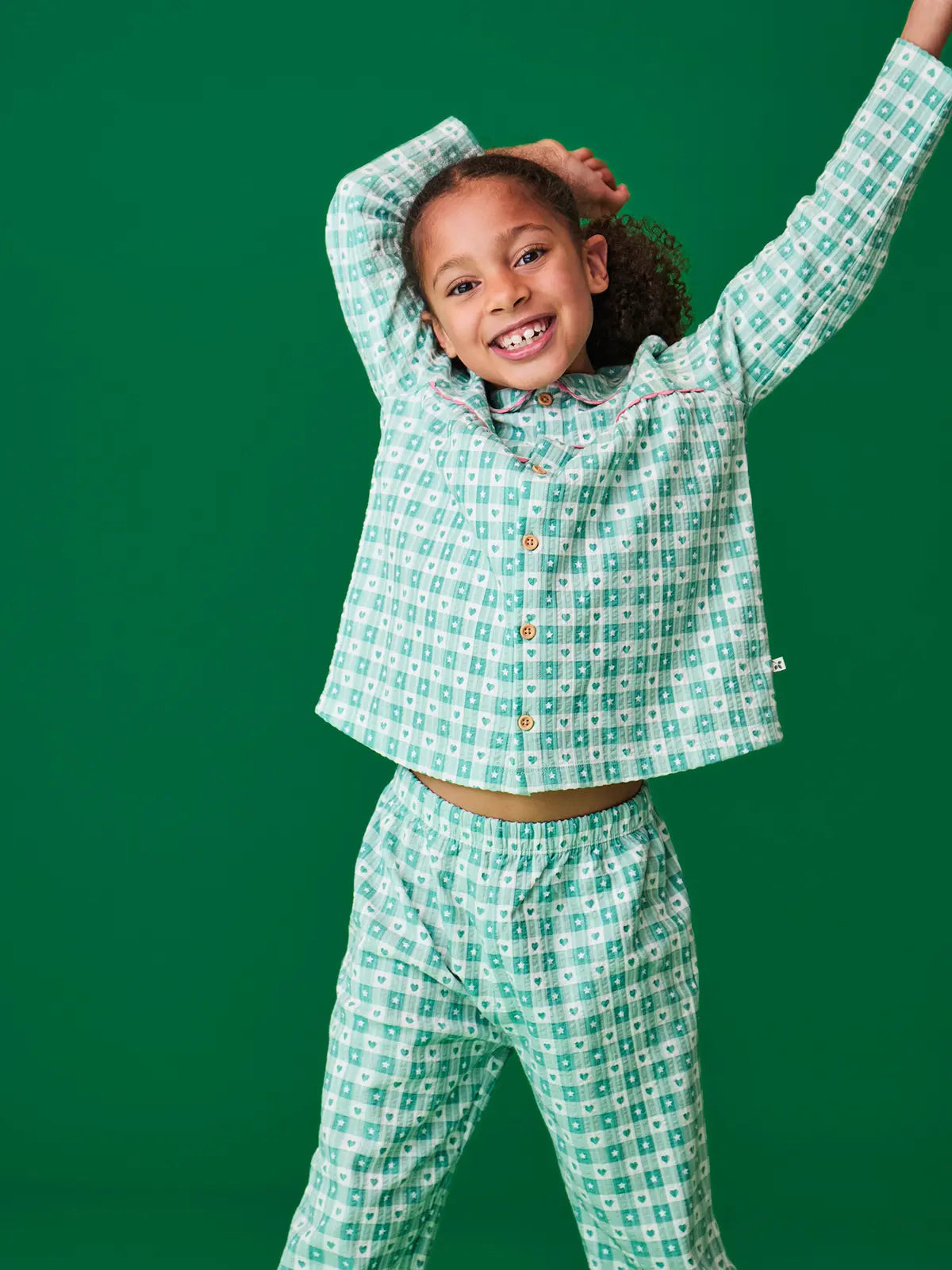 Child wearing the Frugi magic nights organic cotton hayley traditional pyjamas. Light green and white check style with hearts and stars. Pink frill around the collar and across the chest.
