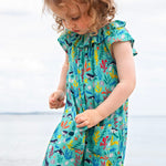 A toddler wearing the Frugi Maris Playsuit, featuring a Sea Life print which includes different sea animals and colourful seaweed and coral, walking on a pebbled beach.