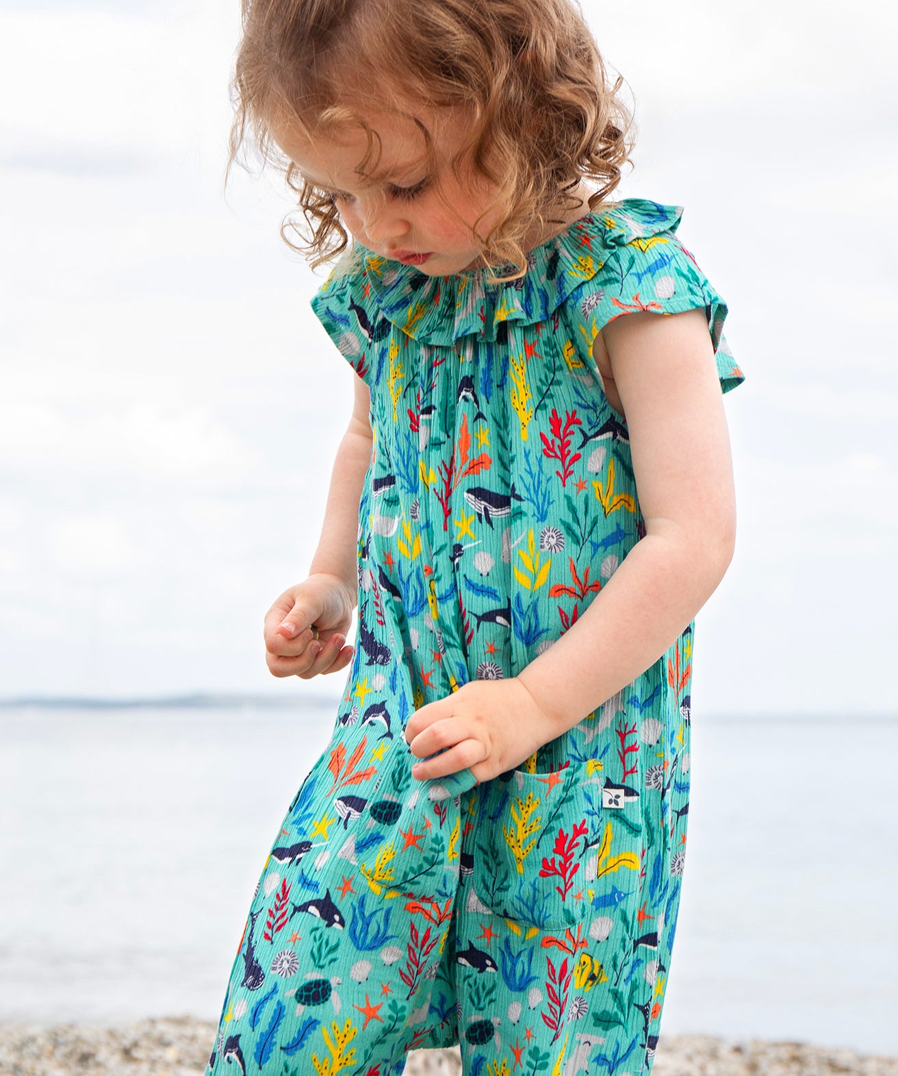 A toddler wearing the Frugi Maris Playsuit, featuring a Sea Life print which includes different sea animals and colourful seaweed and coral, walking on a pebbled beach.