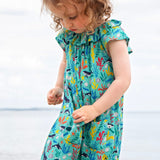 A toddler wearing the Frugi Maris Playsuit, featuring a Sea Life print which includes different sea animals and colourful seaweed and coral, walking on a pebbled beach.