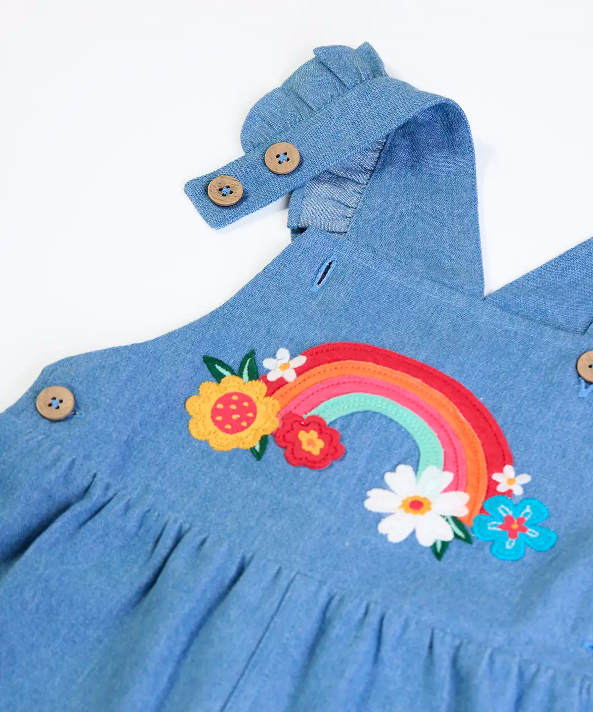 Close up of the rainbow details and button up straps on the Frugi mia denim dungaree with ruffle shoulders and rainbow applique design on the front.