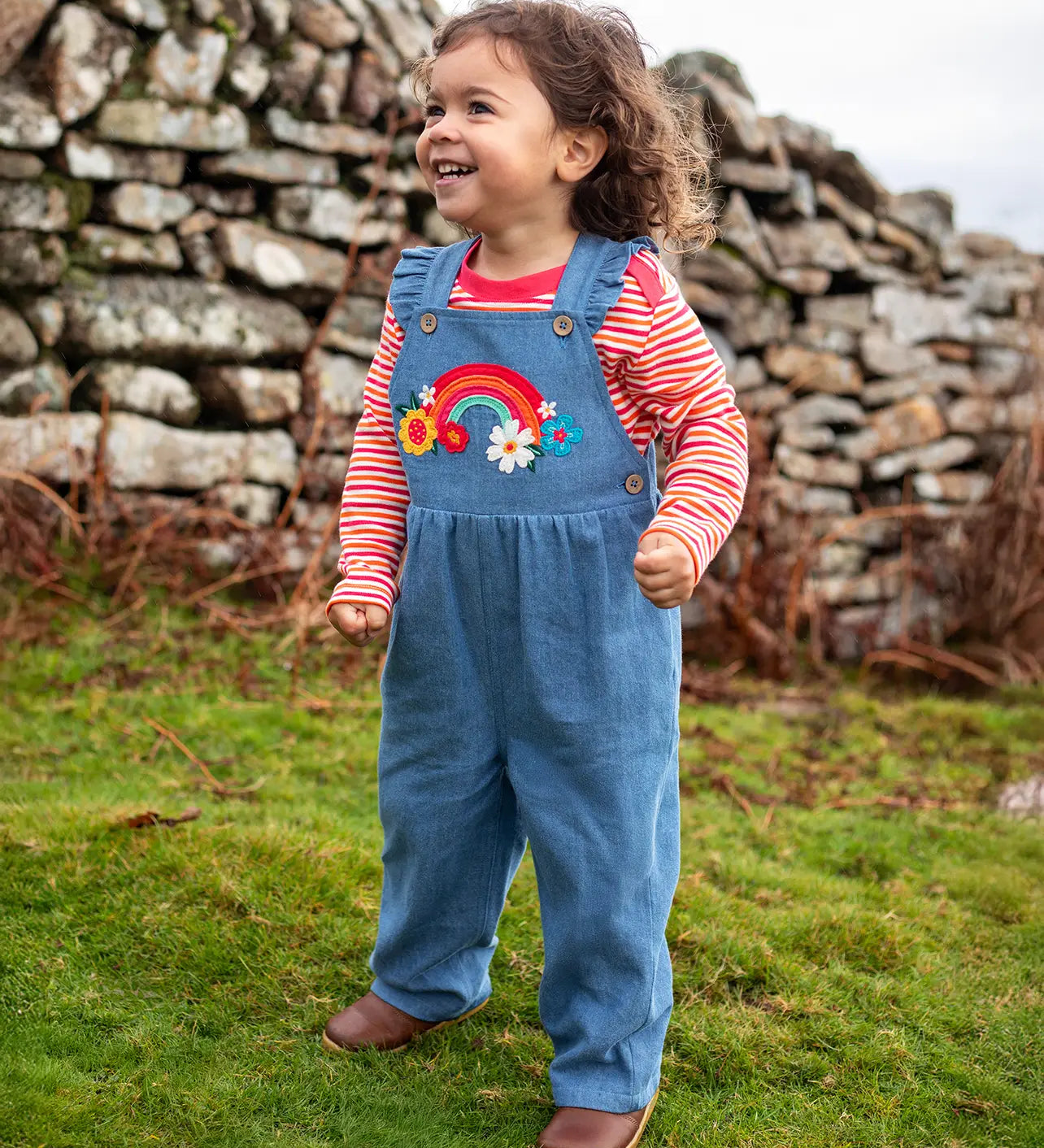 Child wearing the Frugi mia denim dungaree with ruffle shoulders and rainbow applique design on the front with a long sleeved sunset stripe top underneath. 