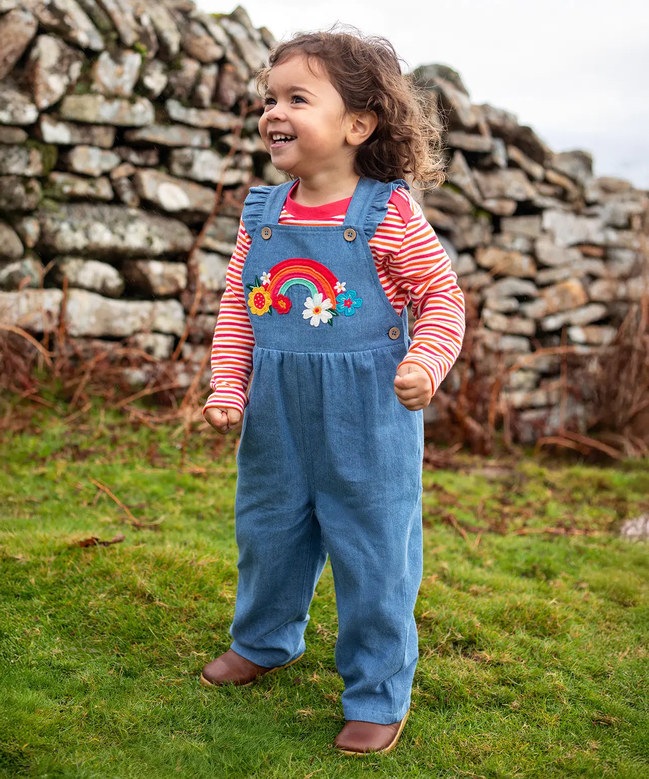 Child wearing the Frugi mia denim dungaree with ruffle shoulders and rainbow applique design on the front with a long sleeved sunset stripe top underneath. 