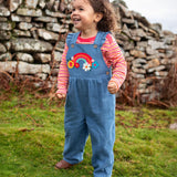 Child wearing the Frugi mia denim dungaree with ruffle shoulders and rainbow applique design on the front with a long sleeved sunset stripe top underneath. 