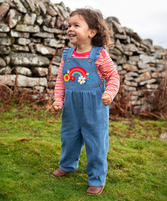 Child wearing the Frugi mia denim dungaree with ruffle shoulders and rainbow applique design on the front with a long sleeved sunset stripe top underneath. 