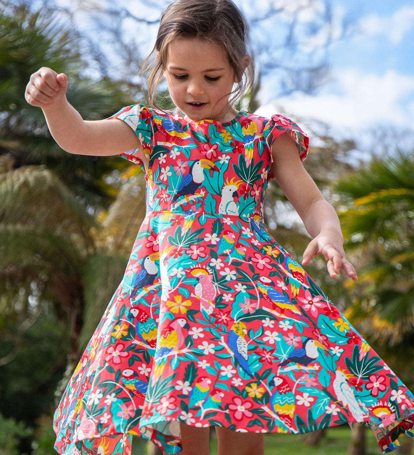 A toddler wearing Frugi Morwenna Skater Dress in the Birds Of Paradise print. Featuring different exotic birds surrounded by colourful flowers and leaves, twirling around amongst palm trees.