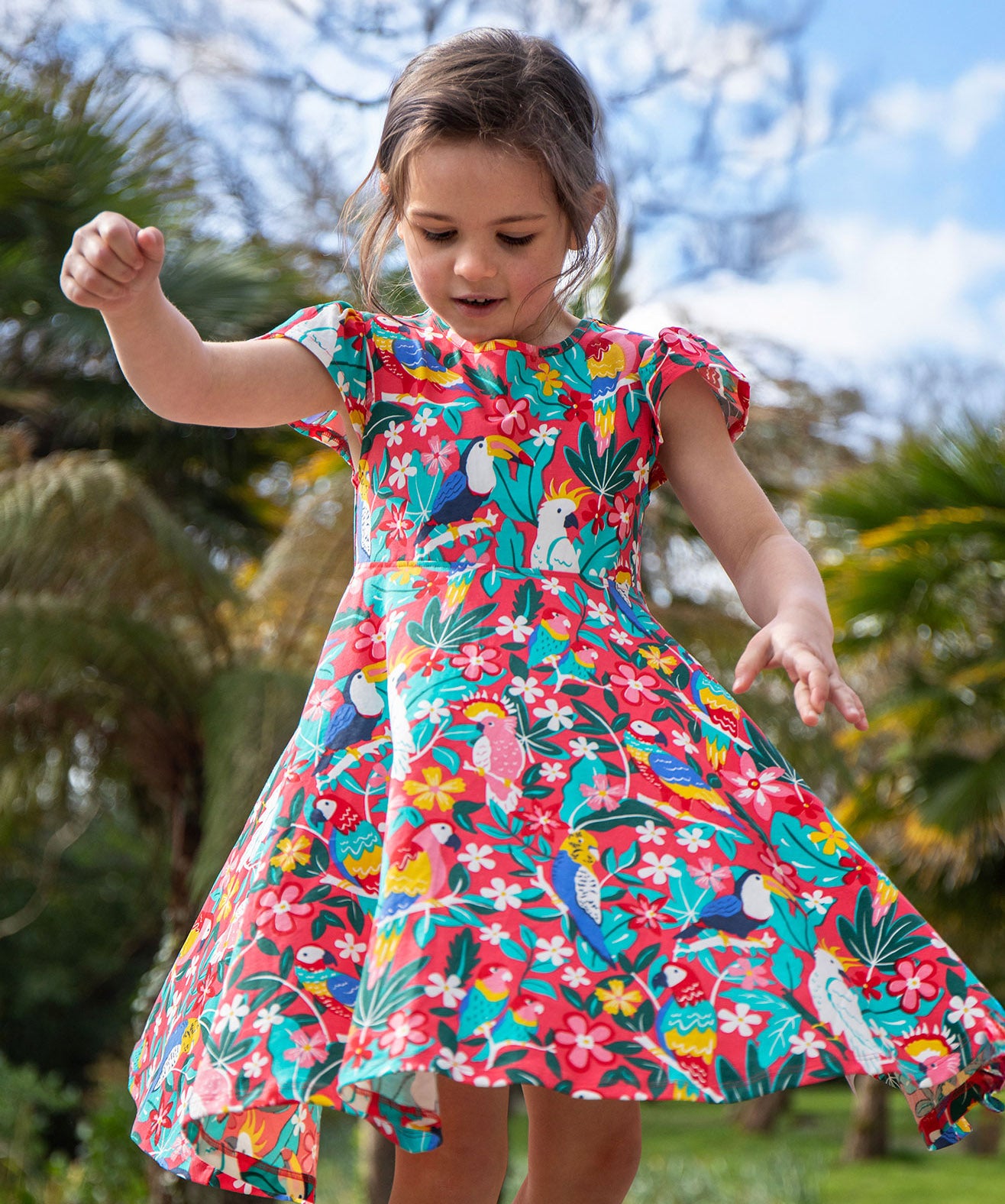 A toddler wearing Frugi Morwenna Skater Dress in the Birds Of Paradise print. Featuring different exotic birds surrounded by colourful flowers and leaves, twirling around amongst palm trees.