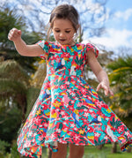 A toddler wearing Frugi Morwenna Skater Dress in the Birds Of Paradise print. Featuring different exotic birds surrounded by colourful flowers and leaves, twirling around amongst palm trees.