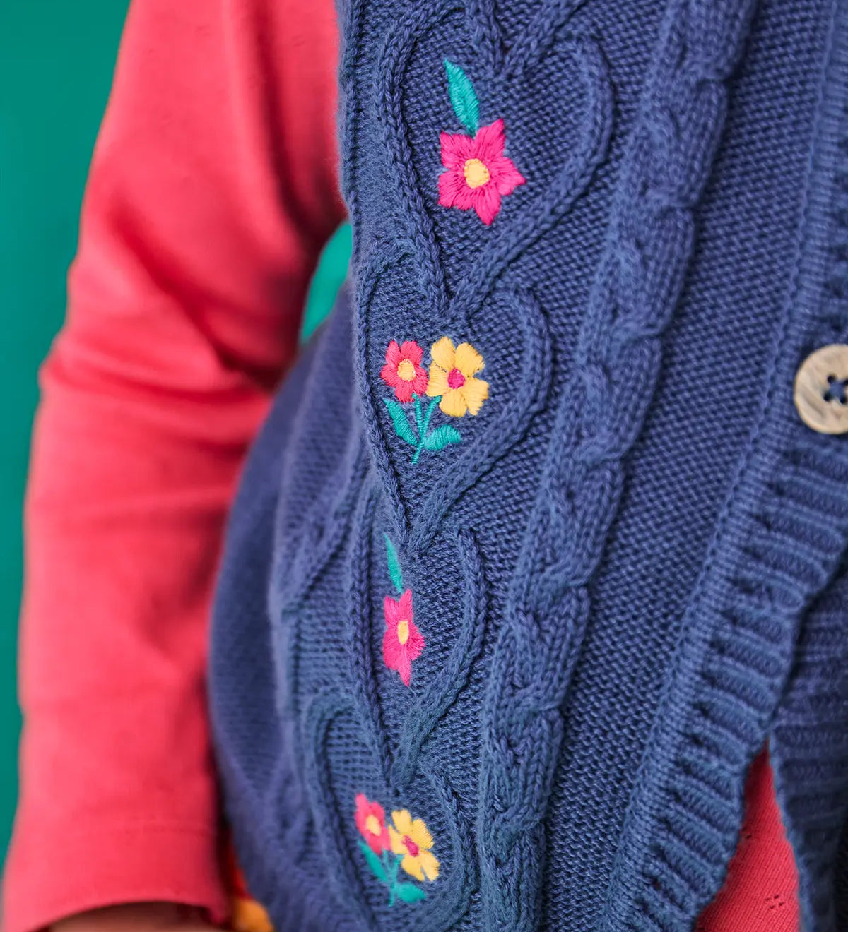 Close up of the Frugi organic cotton connie knitted waistcoat with embroidered flower details, wooden button closure and scalloped edges worn by a child with a long sleeved top underneath.