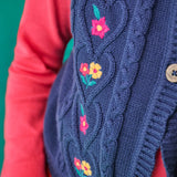 Close up of the Frugi organic cotton connie knitted waistcoat with embroidered flower details, wooden button closure and scalloped edges worn by a child with a long sleeved top underneath.