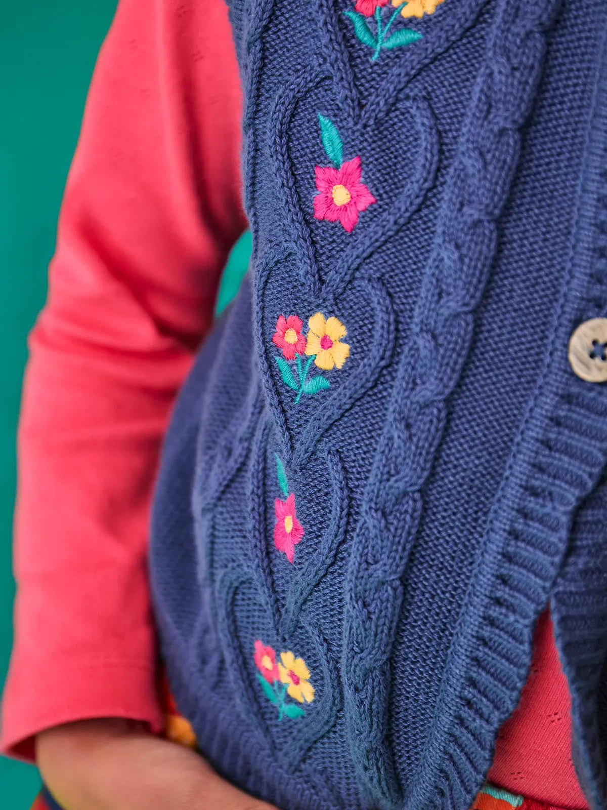 Close up of the Frugi organic cotton connie knitted waistcoat with embroidered flower details, wooden button closure and scalloped edges worn by a child with a long sleeved top underneath.