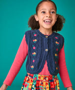 Child wearing the Frugi organic cotton connie navy knitted waistcoat with embroidered flower details, wooden button closure and scalloped edges with a pink long sleeve top underneath