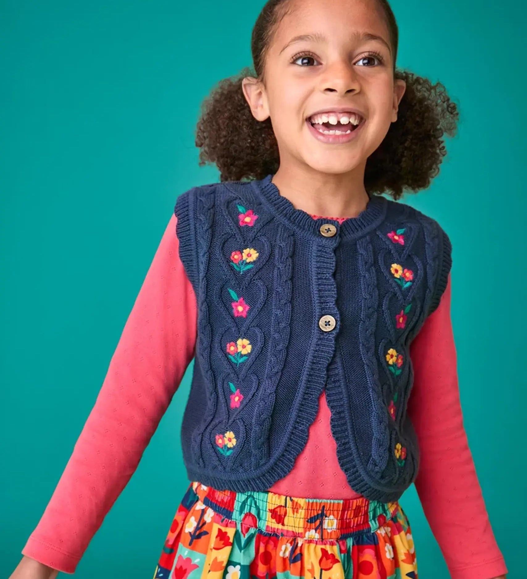 Child wearing the Frugi organic cotton connie navy knitted waistcoat with embroidered flower details, wooden button closure and scalloped edges with a pink long sleeve top underneath