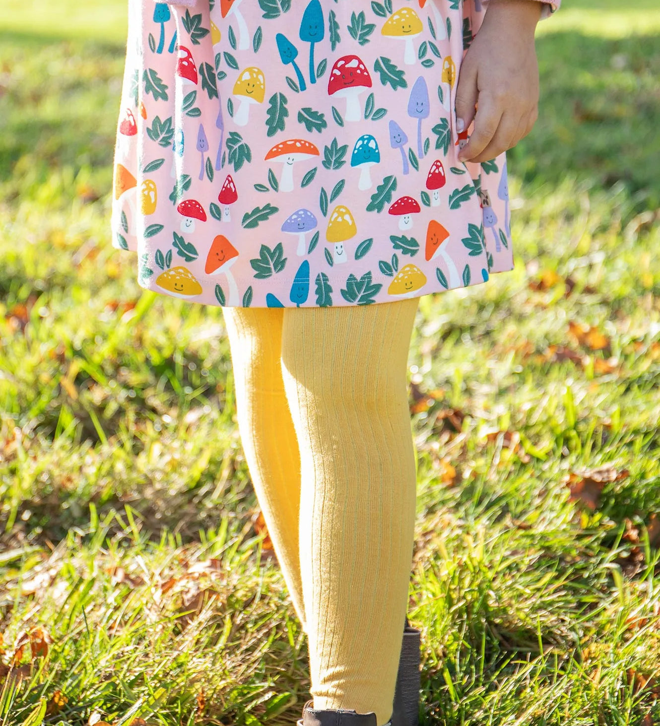 A closeup of a girl's legs wearing the Frugi organic cotton Norah Tights in bumblebee yellow, paired with the Amelia Dress with a mushroom design.