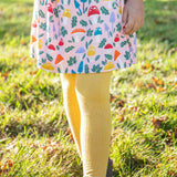 A closeup of a girl's legs wearing the Frugi organic cotton Norah Tights in bumblebee yellow, paired with the Amelia Dress with a mushroom design.