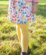 A closeup of a girl's legs wearing the Frugi organic cotton Norah Tights in bumblebee yellow, paired with the Amelia Dress with a mushroom design.