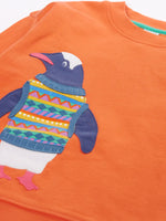 Close up of the Frugi organic cotton superb sweatshirt salamander orange with penguin applique. The penguin is wearing a fairisle style tank top. 
