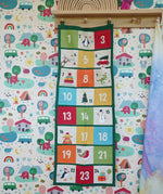 Colourful Frugi forest festive fun organic cotton advent calendar hung up on wooden peg in the Babipur playroom. 24 pockets with alternating colourful illustrations and colour blocks. 