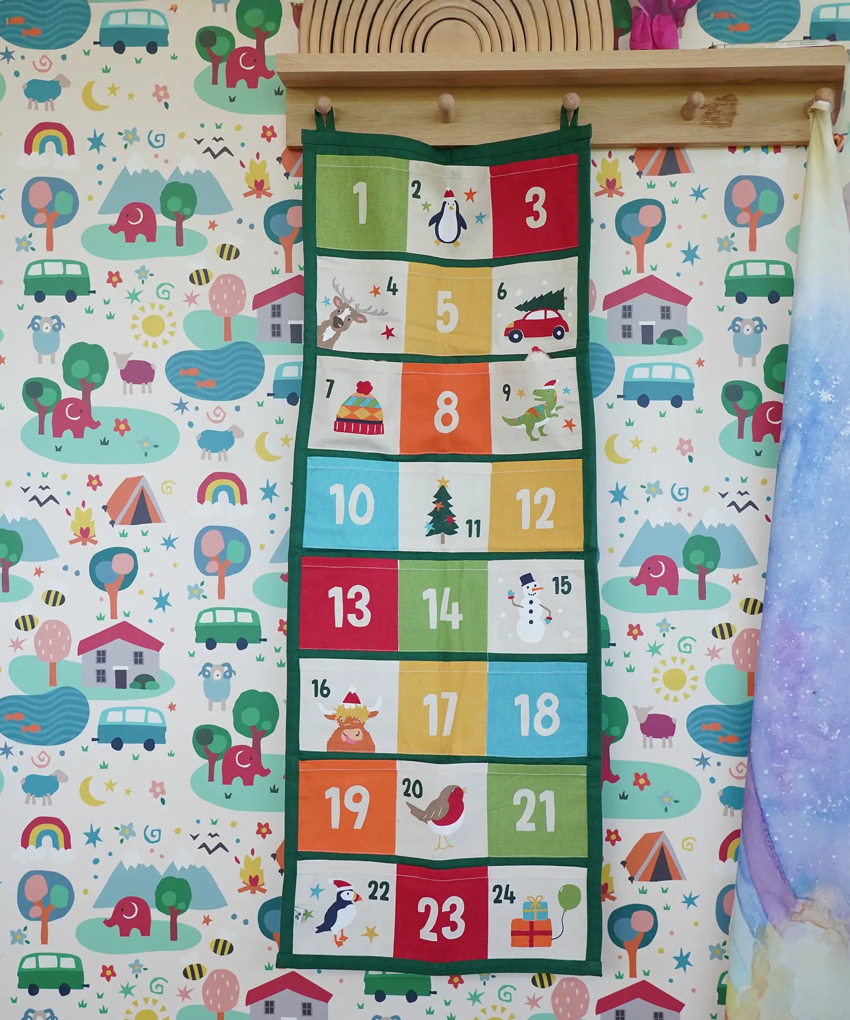 Colourful Frugi forest festive fun organic cotton advent calendar hung up on wooden peg in the Babipur playroom. 24 pockets with alternating colourful illustrations and colour blocks. 