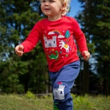 Child wearing the Frugi red organic cotton adventure magic applique top. Long sleeved top with castle, rainbow, tree horse and nessie appliques on the front. 