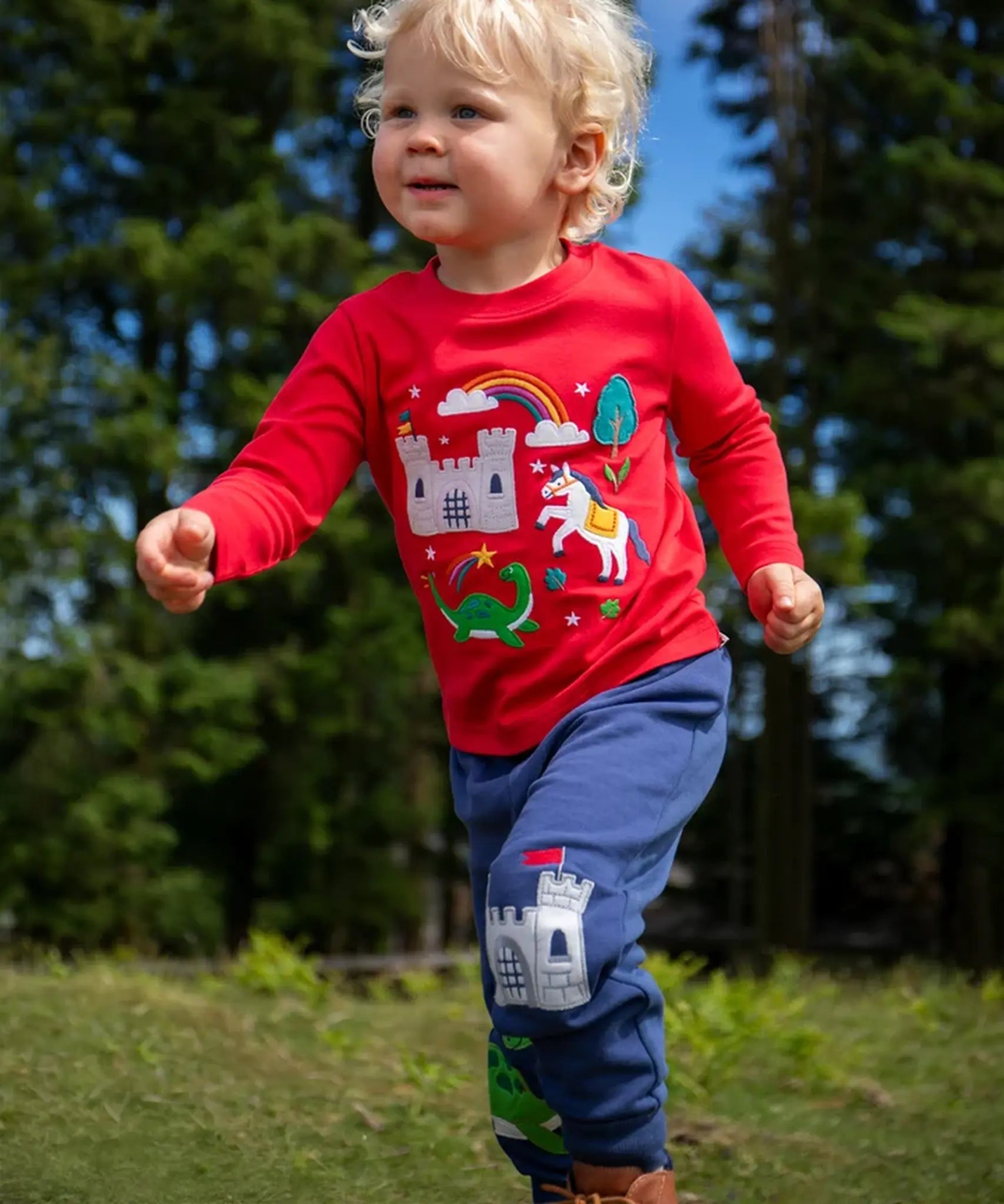 Child wearing the Frugi red organic cotton adventure magic applique top. Long sleeved top with castle, rainbow, tree horse and nessie appliques on the front. 
