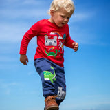 Child wearing the Frugi red organic cotton adventure magic applique top. Long sleeved top with castle, rainbow, tree horse and nessie appliques on the front. 