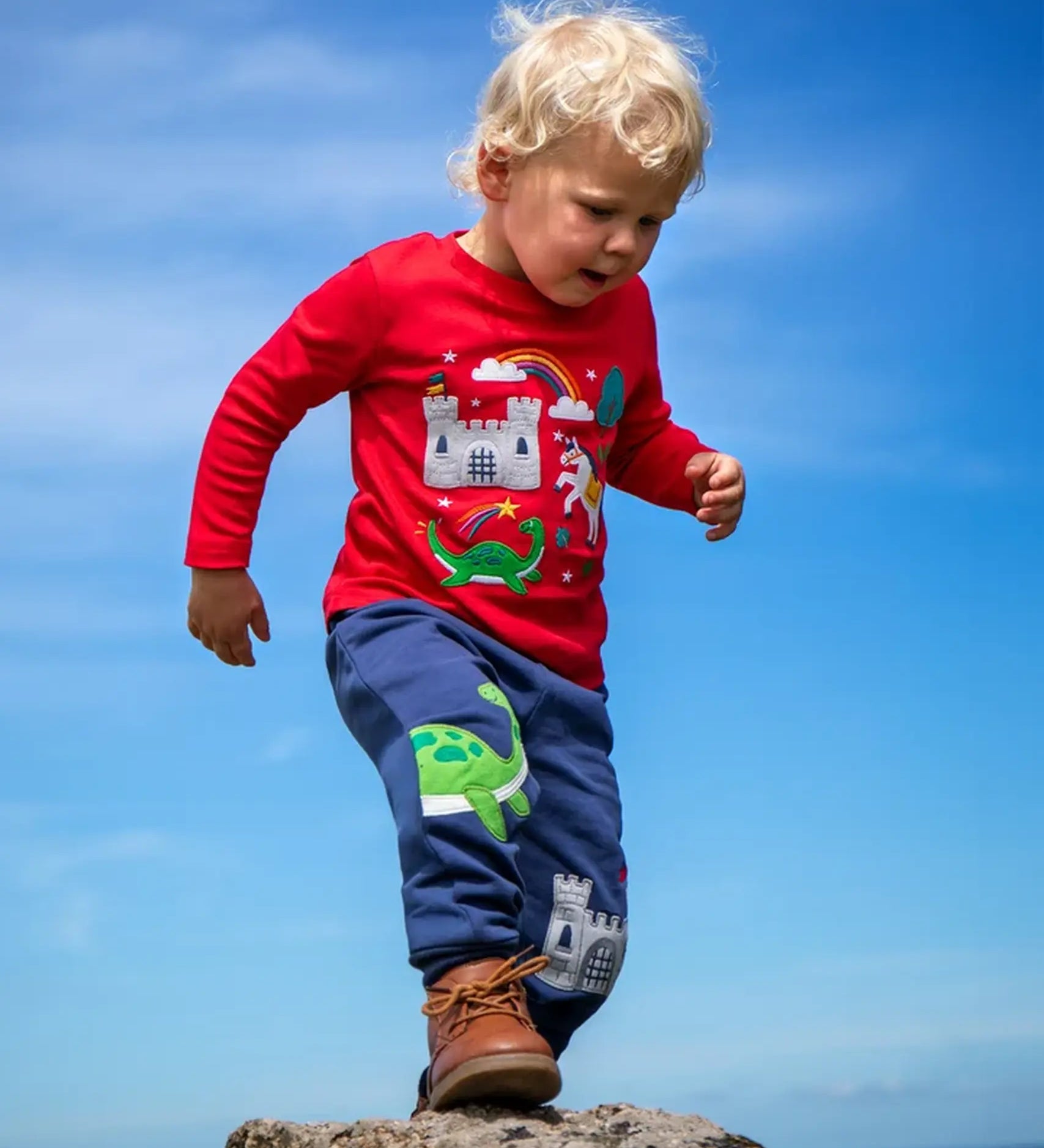 Child wearing the Frugi red organic cotton adventure magic applique top. Long sleeved top with castle, rainbow, tree horse and nessie appliques on the front. 