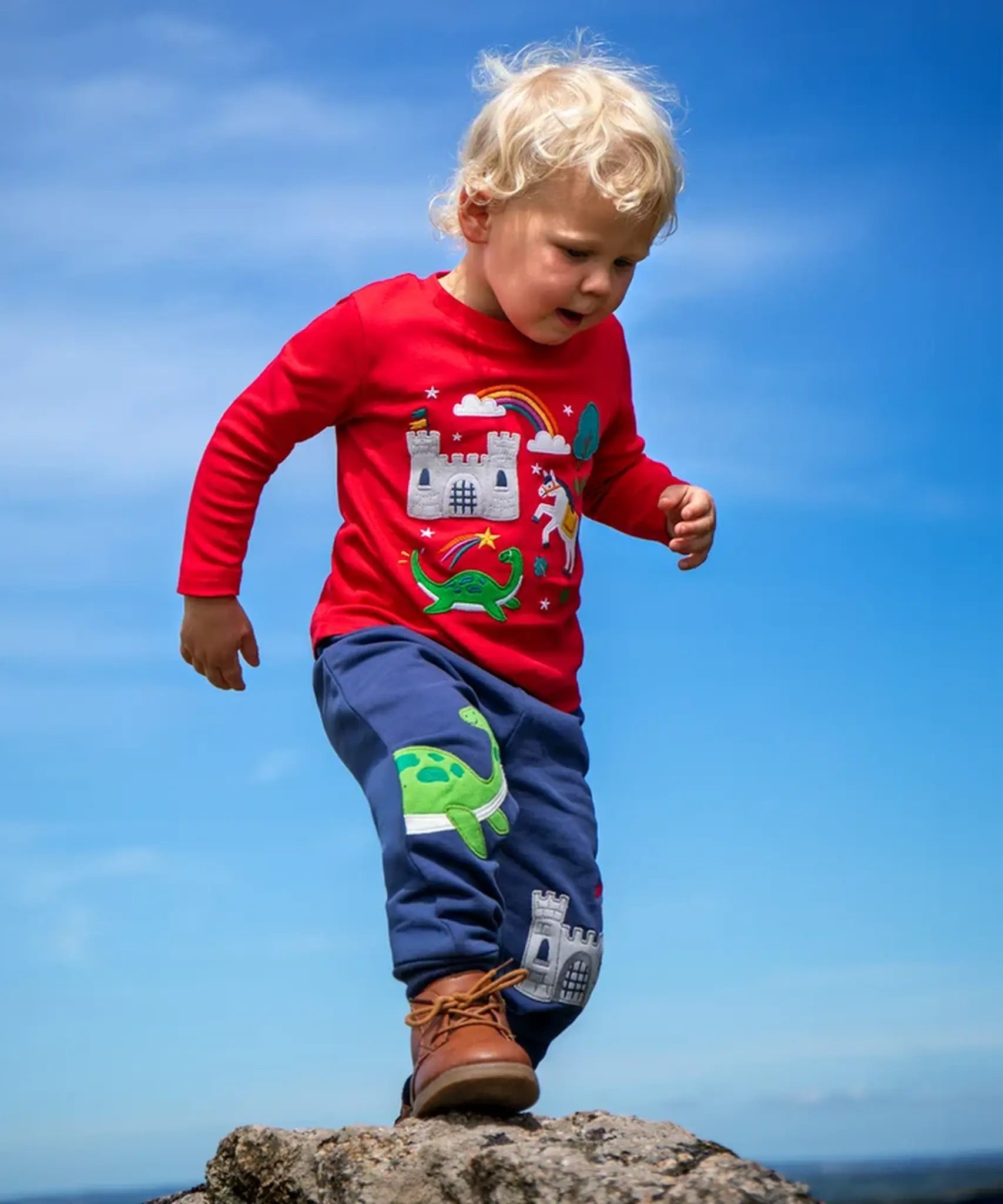 Child wearing the Frugi red organic cotton adventure magic applique top. Long sleeved top with castle, rainbow, tree horse and nessie appliques on the front. 