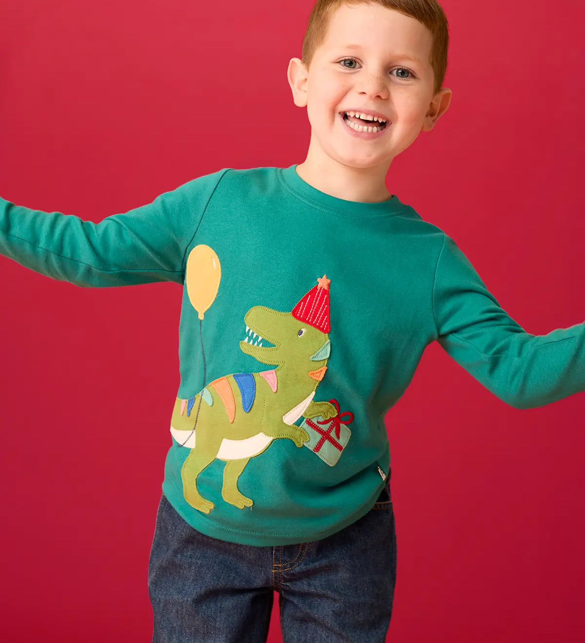Child wearing the Frugi organic cotton adventure long sleeve hut green child's top with t-rex dinosaur wearing a party hat carrying a present with a yellow balloon appliques on the front with denim jeans.