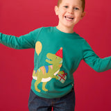 Child wearing the Frugi organic cotton adventure long sleeve hut green child's top with t-rex dinosaur wearing a party hat carrying a present with a yellow balloon appliques on the front with denim jeans.