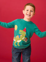Child wearing the Frugi organic cotton adventure long sleeve hut green child's top with t-rex dinosaur wearing a party hat carrying a present with a yellow balloon appliques on the front with denim jeans.