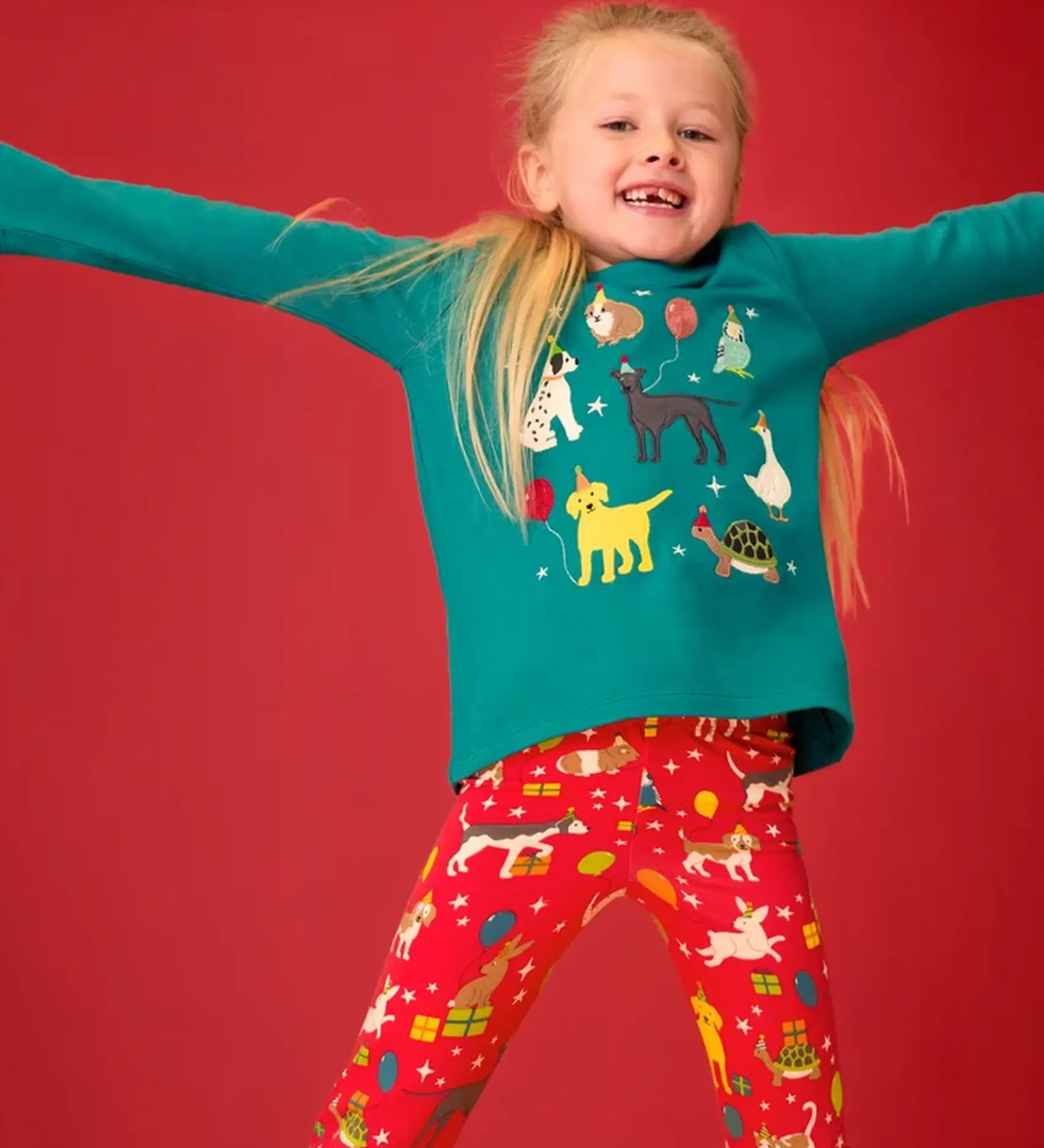 Child wearing the Frugi organic cotton anna pet Paw-Ty appliqué long sleeve children's top with red matching print leggings. Green top with applique dogs, guinea pig, budgie, goose and tortoise wearing party hats and balloons and star sparkle details. 