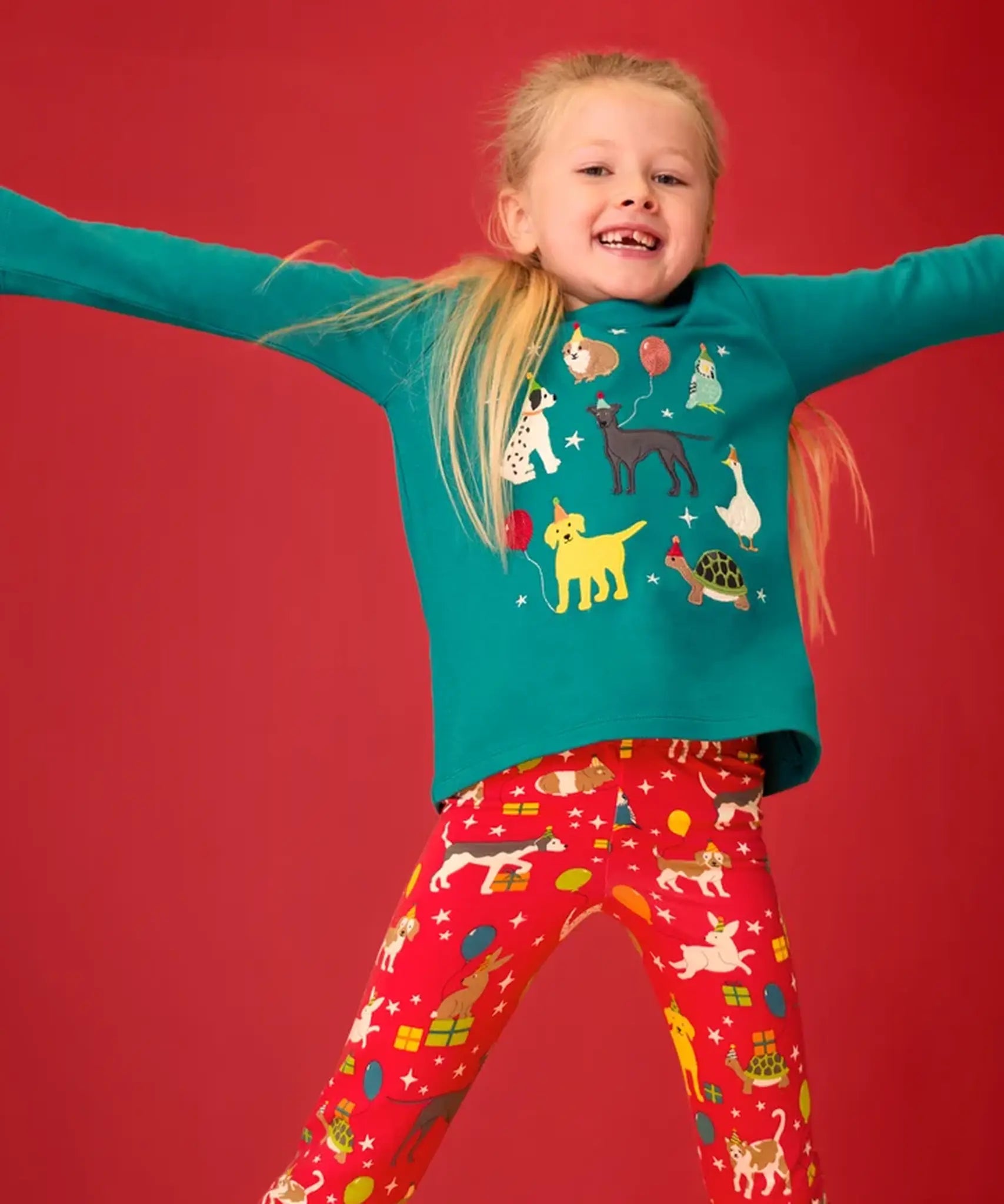 Child wearing the Frugi organic cotton anna pet Paw-Ty appliqué long sleeve children's top with red matching print leggings. Green top with applique dogs, guinea pig, budgie, goose and tortoise wearing party hats and balloons and star sparkle details. 