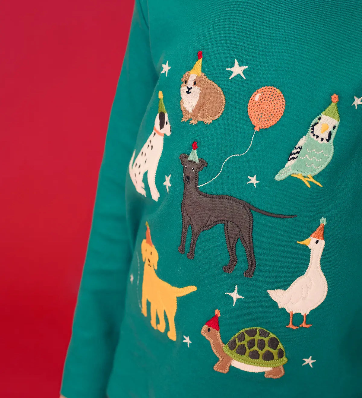 Close up of child wearing the Frugi organic cotton anna pet Paw-Ty appliqué long sleeve children's top. Green top with applique dogs, guinea pig, budgie, goose and tortoise wearing party hats and balloons and star sparkle details. 