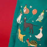 Close up of child wearing the Frugi organic cotton anna pet Paw-Ty appliqué long sleeve children's top. Green top with applique dogs, guinea pig, budgie, goose and tortoise wearing party hats and balloons and star sparkle details. 