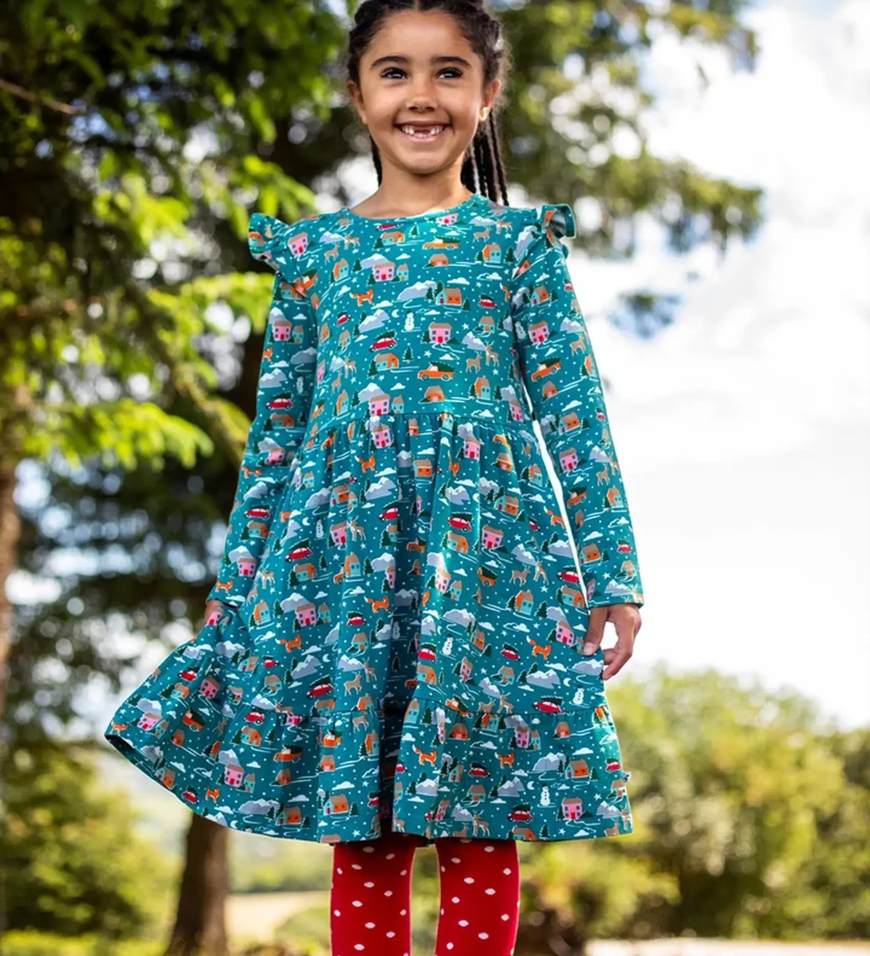 Child wearing the Frugi organic cotton annika skater dress with winter drives print. All over print featuring houses, campers, snow topped mountains, snowman and trees in a festive scene. 