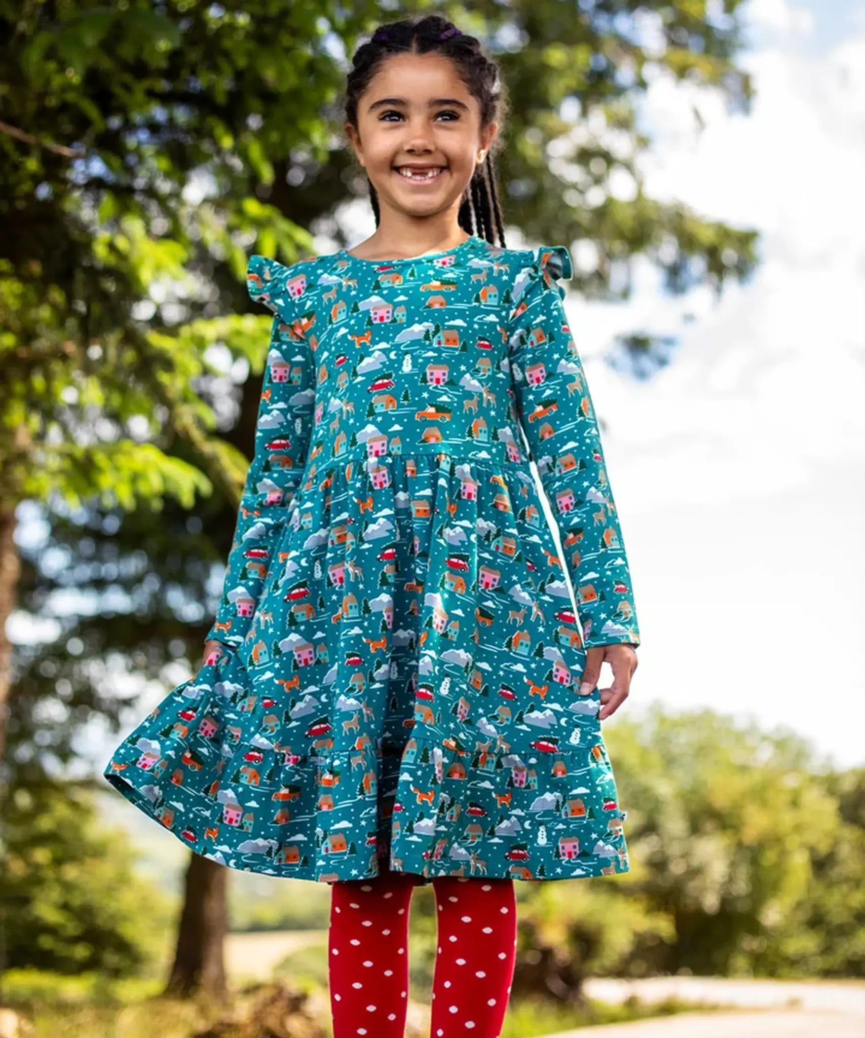 Child wearing the Frugi organic cotton annika skater dress with winter drives print. All over print featuring houses, campers, snow topped mountains, snowman and trees in a festive scene. 