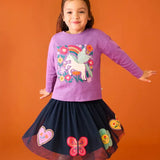 Child wearing the Frugi organic cotton Bethany long sleeved periwinkle purple top with a pegasus applique with a rainbow and flowers with a navy skirt. 