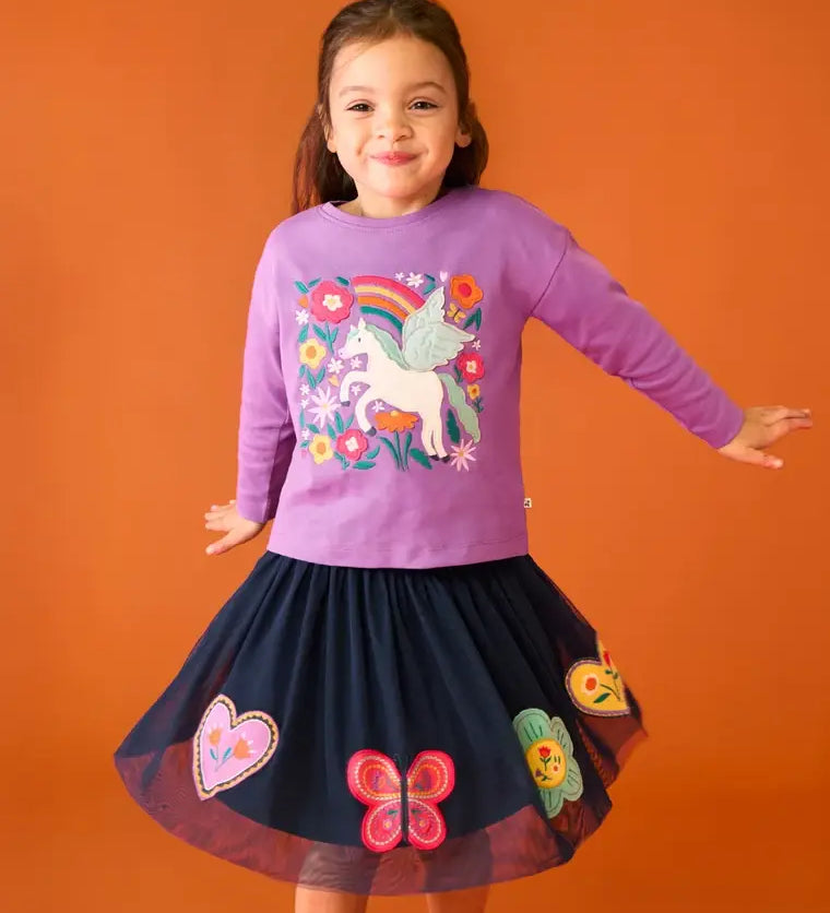 Child wearing the Frugi organic cotton Bethany long sleeved periwinkle purple top with a pegasus applique with a rainbow and flowers with a navy skirt. 