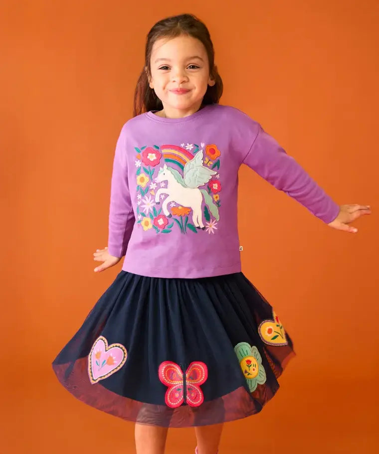 Child wearing the Frugi organic cotton Bethany long sleeved periwinkle purple top with a pegasus applique with a rainbow and flowers with a navy skirt. 