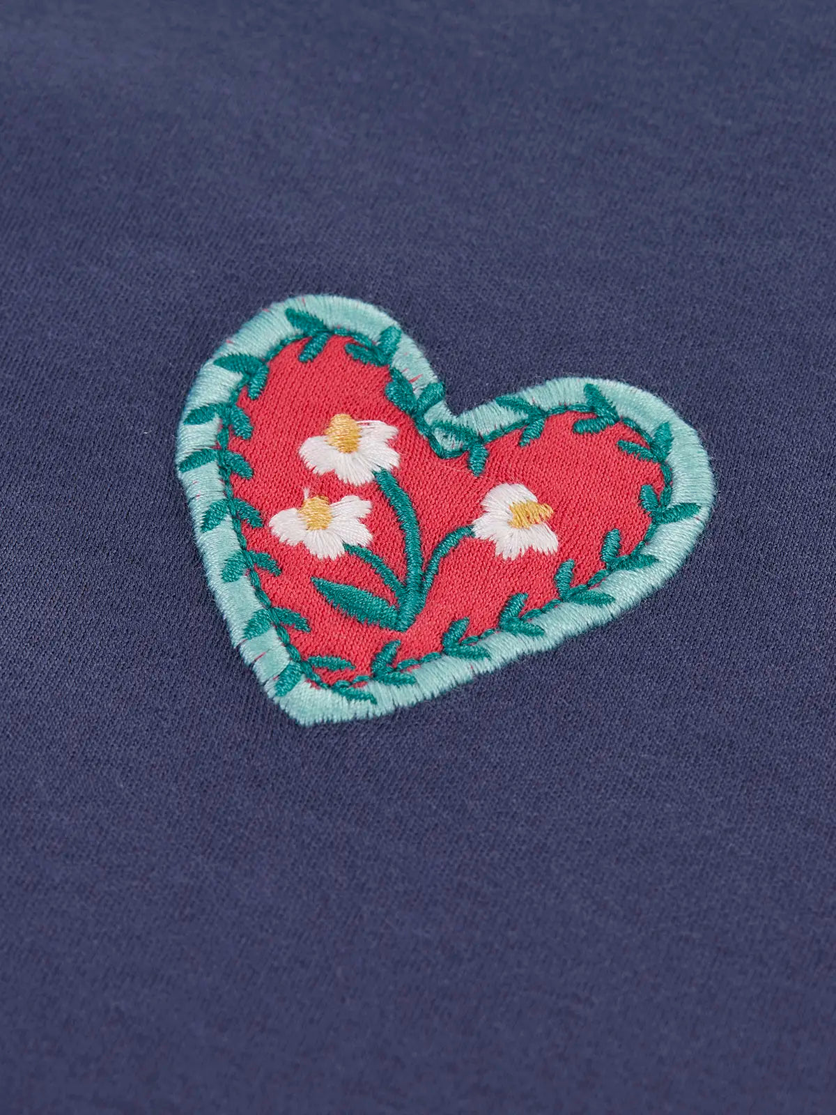 Close up of a heart applique featured on the Frugi bethany organic cotton long sleeved child's navy top with floral embroidery details.