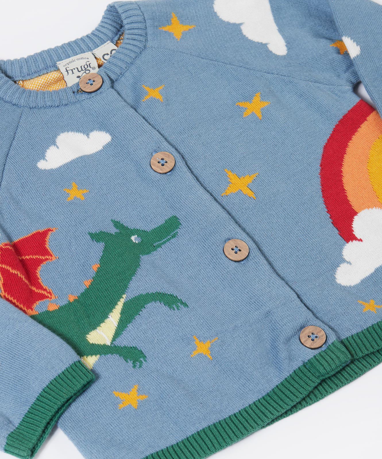 A close up of the front of the Frugi Organic Cotton Reva Dragon Skies Knitted Kids' Cardigan. This blue cardigan features a fun, colourful design featuring a green dragon, stars, rainbow, clouds and a mushroom. This cardigan is a part of a wide range of organic cotton clothing for children available here at Babipur. 