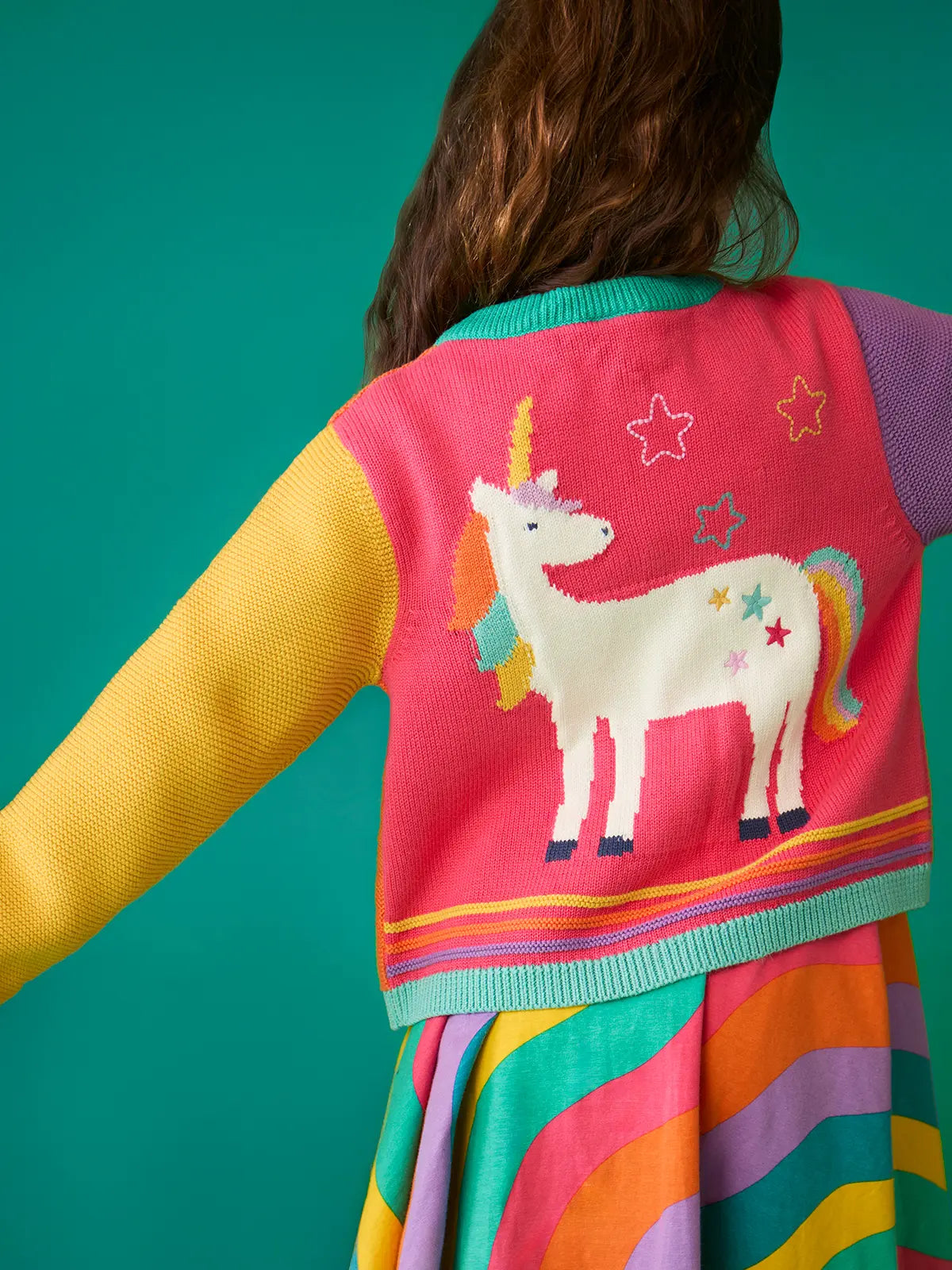 Back of a child wearing the Frugi organic cotton cammie cardigan showing the large unicorn on the back with a rainbow dress underneath.