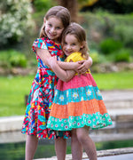 Two children hugging, one is wearing the Frugi Children's  Rainbow Hotchpotch Organic Dorothy Twirly Dress and the other is wearing the Frugi  Birds Of Paradise Morwenna Skater Dress .