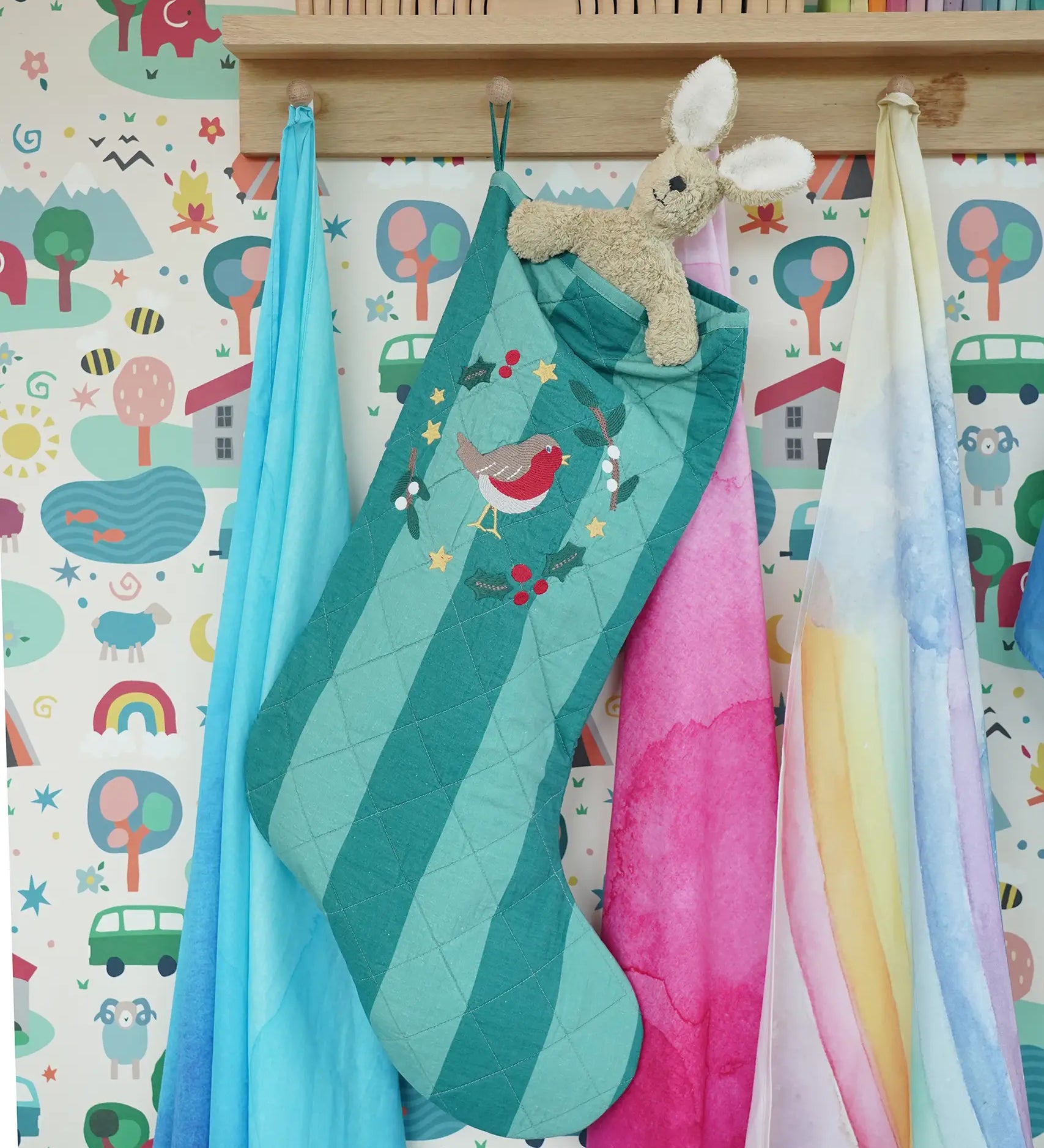 Frugi organic cotton Christmas stocking. Striped teal quilted style stocking with a robin wreath embroidery detail and hanging loop hung up in the Babipur playroom with a Senger rabbit soft toy inside.